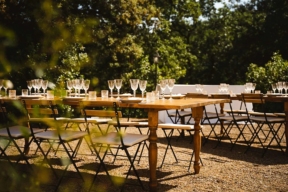Long wooden tables laid with glasses outdoors under the trees