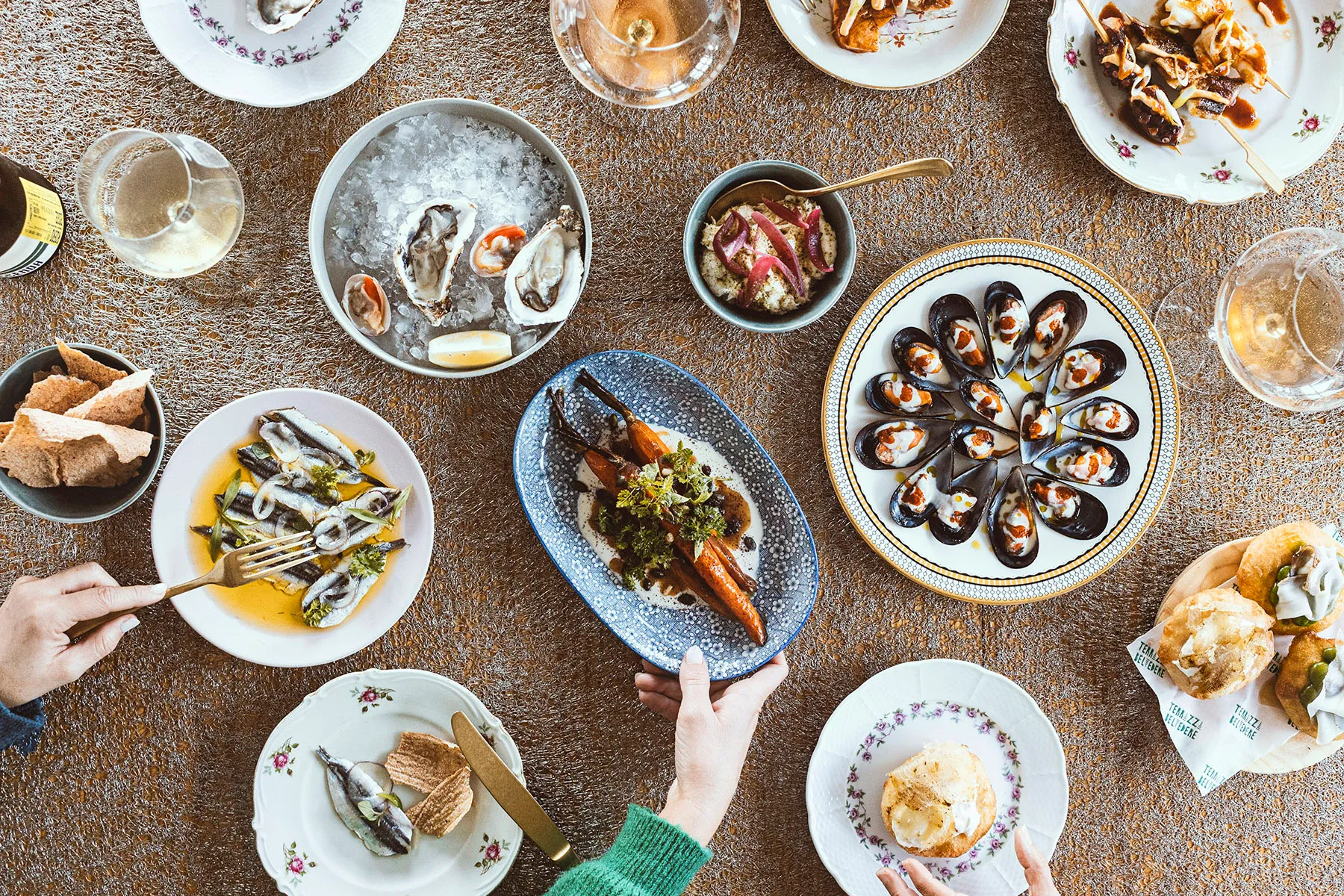 Overhead view of a shared table with oysters, mussels, sardines and wine, hands reaching for the plates